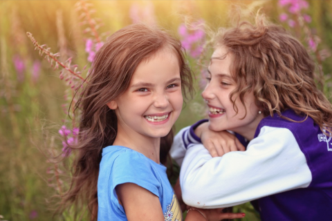 Two young girls smile while standing in a field of tall grass.