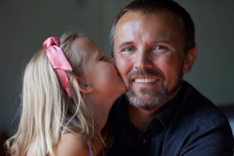 Daughter with a pink bow on her head kisses the cheek of her smiling father