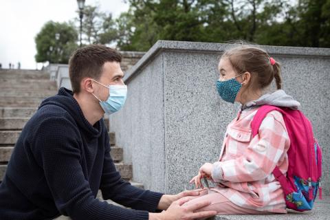 A young girl wearing a face mask and a pink backpack sits on a ledge with her dad hold her knees.
