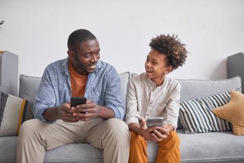 A father and son sit on a couch together as they both use their smartphones.