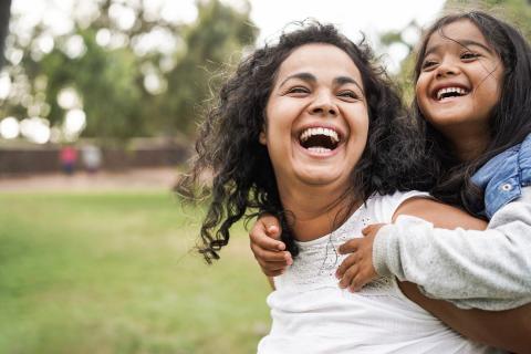 A happy mother gives her smiling daughter a piggy ride in the park.