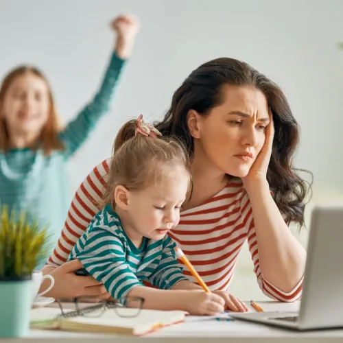 A busy mother looks worried at her computer as her children play around her.
