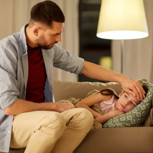 Father checks daughter's temperature as she lays on the couch