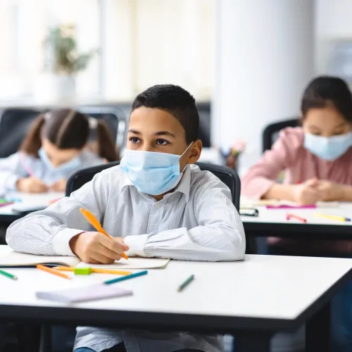 A young boy wears a face mask looks up as he works at a desk in a classroom filled with kids also wearing face masks.
