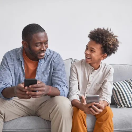 A father and son sit on a couch together as they both use their smartphones.