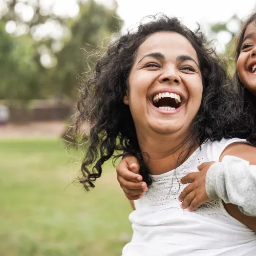 A happy mother gives her smiling daughter a piggy ride in the park.