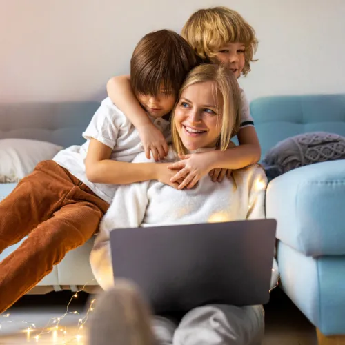 A mother works on a computer while her two sons hug her from behind.