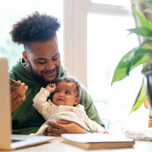 A father holds his infant while working on a computer.