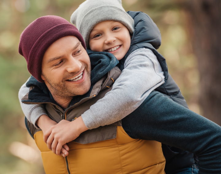 A father gives his young son a piggy back ride while on a walk in the woods.