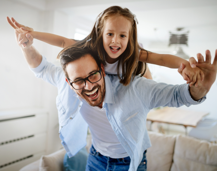 A father carries his young daughter on his back. They both hold their arms out wide.