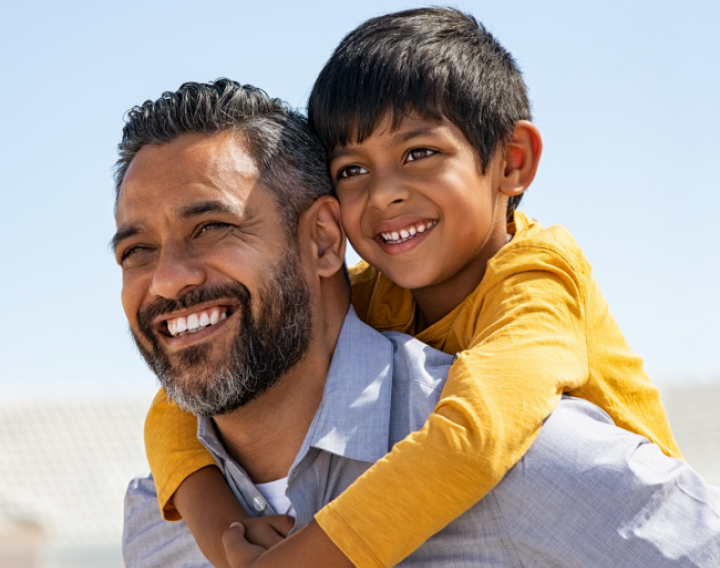 Bearded father in blue button up gives his son, who is wearing a yellow shirt, a piggy back ride outside