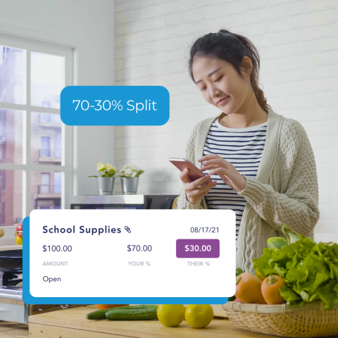 A woman stands in a kitchen and looks down at her smartphone to review an expense for school supplies.