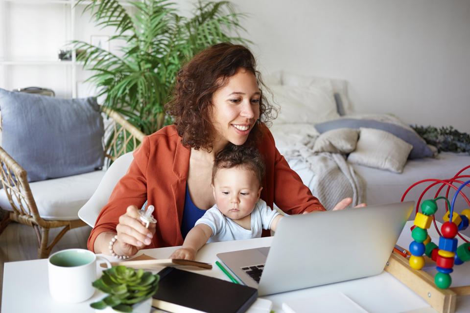 Mother works on laptop as she holds her young child in her lap.