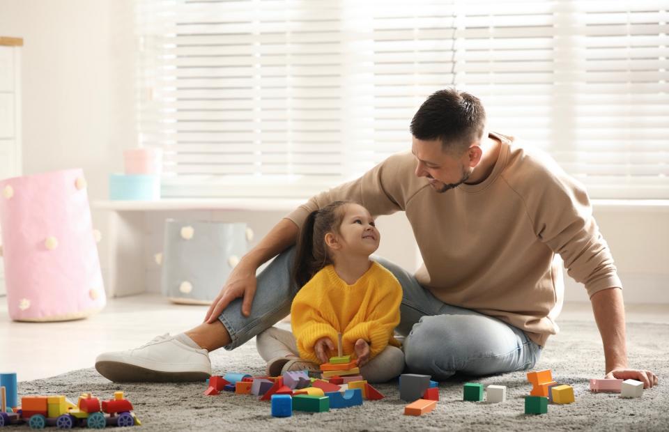 A father and daughter sit together on the floor to play with blocks and smile at each other.