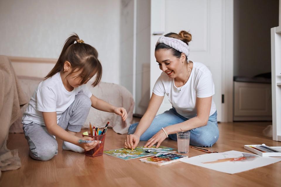 Mother and daughter sit on the floor together to paint pictures on paper.