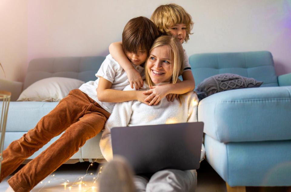 A mother works on a computer while her two sons hug her from behind.